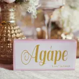 Cartel rosa con la palabra "Agápe" en dorado, junto a un jarrón dorado y flores.