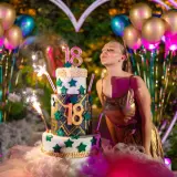 Joven soplando velas en un pastel de tres pisos para celebrar su 18 cumpleaños, rodeada de globos y luces.