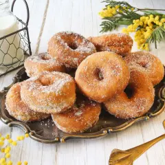 Un plato de rosquillas caseras de toda la vida, espolvoreadas con azúcar, junto a botellas de leche y flores amarillas.