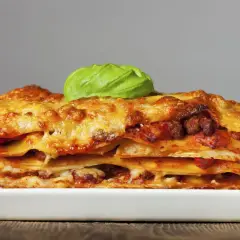 Una porción generosa de lasaña de carne, con capas de pasta, salsa y queso dorado, coronada con una hoja de albahaca fresca.