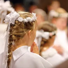 Niña con corona de flores y trenza adornada, rezando en una ceremonia de comunión.