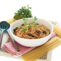 Un tazón de espagueti con salsa putanesca, aceitunas negras y perejil fresco, acompañado de pasta cruda y utensilios de cocina.