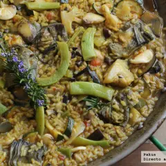 Paella de verduras recién hecha, rebosante de alcachofas, calabacín, pimientos y coliflor, con una ramita de romero y flores moradas.