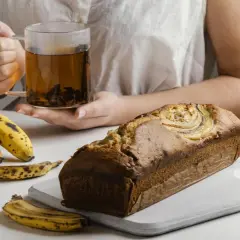 Un delicioso banana bread recién horneado, acompañado de plátanos maduros, nueces y una taza de té humeante.