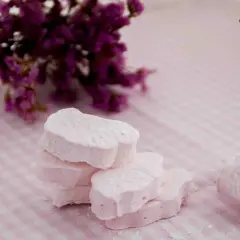 Delicados nubes dulces rosados, espolvoreados con azúcar glas, junto a un ramo de flores moradas sobre un mantel de cuadros.