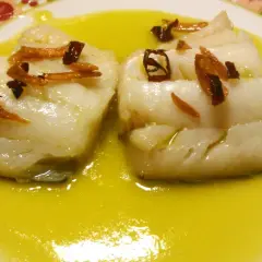 Dos trozos de bacalao al pil pil, cubiertos con aceite de oliva y adornados con ajo frito y guindilla.
