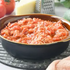 Un tazón de pasta amatriciana, con tomates frescos, queso y rallador al fondo.