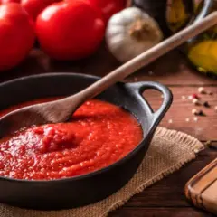Un tazón de tomate frito casero con una cuchara de madera, rodeado de tomates frescos, ajo y aceite de oliva.