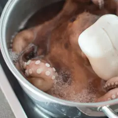 Un pulpo entero se está cociendo en una olla de metal. Sus tentáculos rosados con ventosas visibles se curvan en el agua burbujeante.