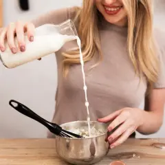 Mujer rubia sonríe mientras vierte leche de una botella a un bol con batidor, preparando algo delicioso.