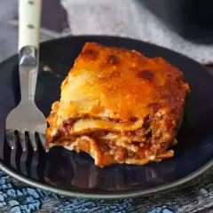 Un trozo de lasaña boloñesa, con capas de pasta, carne y salsa, servido en un plato azul con un tenedor.