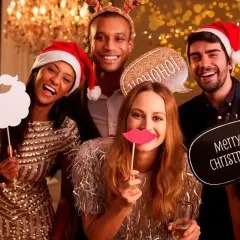 Grupo de amigos sonriendo y riendo en un photocall navideño casero, con accesorios como gafas, barba, labios y carteles de "Ho ho ho" y "Merry Christmas".