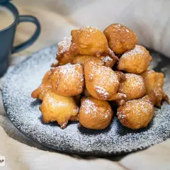 Montón de buñuelos de manzana dorados y espolvoreados con azúcar glas, junto a una taza de café.