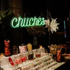 Un letrero de neón verde "Chuchas" ilumina un candy bar lleno de frascos de dulces, piruletas y golosinas.