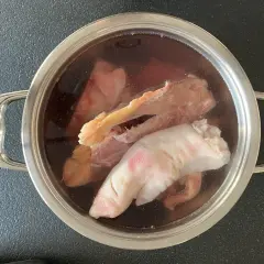 Olla con ingredientes para caldo de huesos: patas de cerdo, huesos y trozos de carne en un líquido oscuro.