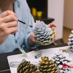 Manos pintando piñas para navidad con pintura plateada y detalles dorados. Decoración festiva en proceso.