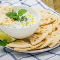 Un cuenco de cremosa salsa tzatziki, decorado con hierbas frescas y un chorrito de aceite, servido con pan de pita crujiente y rodajas de lima.