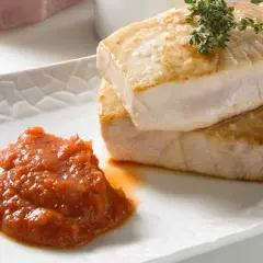 Dos trozos de atún a la plancha, dorados y jugosos, apilados sobre un plato blanco con una porción de salsa de tomate y una ramita de tomillo.