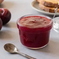 Un tarro de mermelada de ciruela casera, con ciruelas frescas y tostadas al fondo. ¡Perfecta para el desayuno!