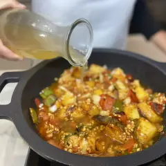 Se vierte caldo sobre un sofrito para paella con pimientos, tomate y patatas en una paellera.