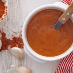 Un cuenco blanco con salsa brava, pimentón en polvo, ajos y una servilleta a rayas rojas.