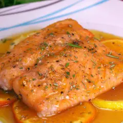 Filete de salmón a la naranja, glaseado y con hierbas, servido sobre rodajas de naranja y limón en una salsa dorada.