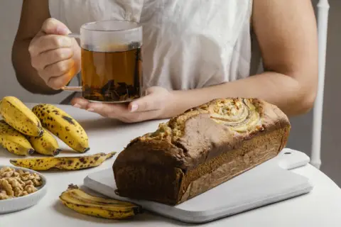Un delicioso banana bread recién horneado, acompañado de plátanos maduros, nueces y una taza de té humeante.
