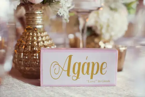 Cartel rosa con la palabra "Agápe" en dorado, junto a un jarrón dorado y flores.
