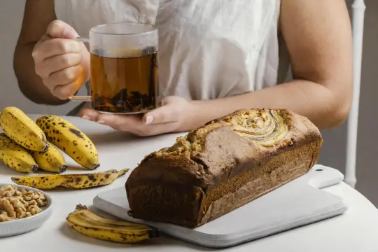 Un delicioso banana bread recién horneado, acompañado de plátanos maduros, nueces y una taza de té humeante.