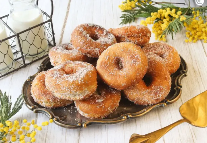Un plato de rosquillas caseras de toda la vida, espolvoreadas con azúcar, junto a botellas de leche y flores amarillas.