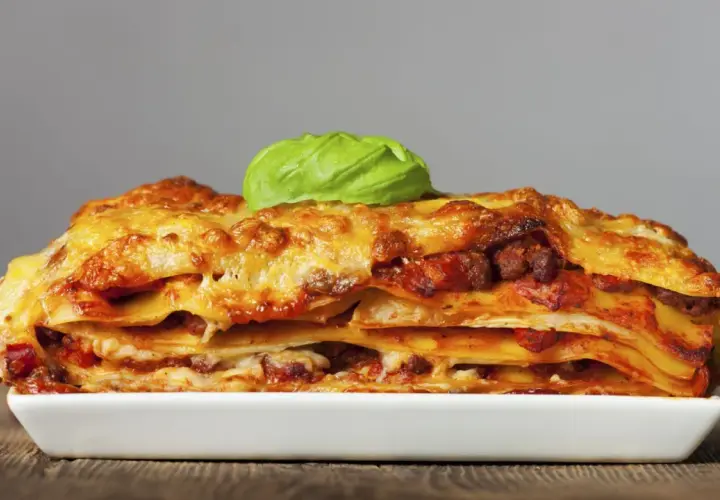 Una porción generosa de lasaña de carne, con capas de pasta, salsa y queso dorado, coronada con una hoja de albahaca fresca.