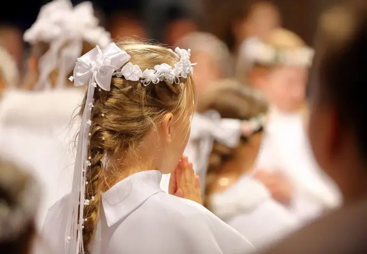 Niña con corona de flores y trenza adornada, rezando en una ceremonia de comunión.