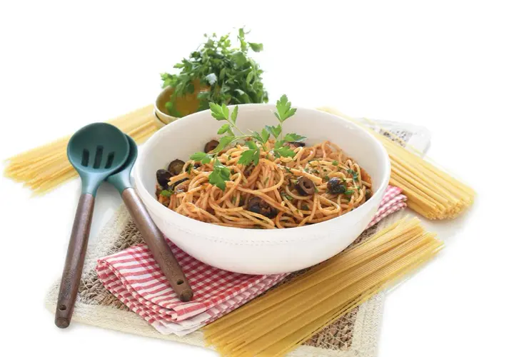 Un tazón de espagueti con salsa putanesca, aceitunas negras y perejil fresco, acompañado de pasta cruda y utensilios de cocina.