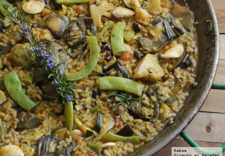 Paella de verduras recién hecha, rebosante de alcachofas, calabacín, pimientos y coliflor, con una ramita de romero y flores moradas.