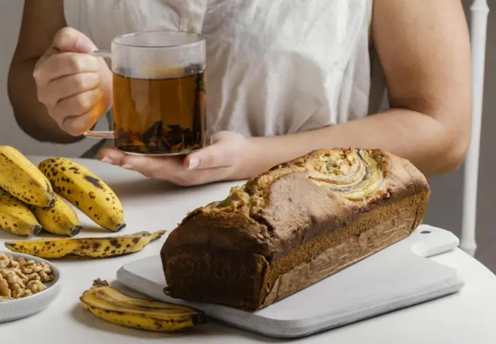 Un delicioso banana bread recién horneado, acompañado de plátanos maduros, nueces y una taza de té humeante.