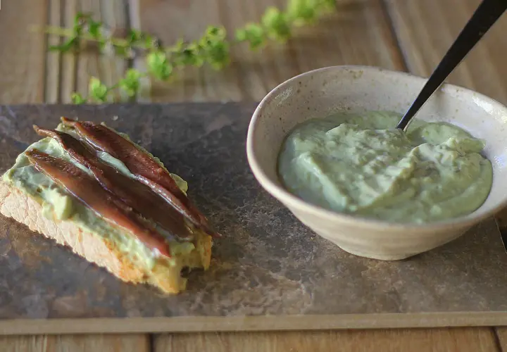 Tosta con mayonesa de aguacate y anchoas. Un cuenco con la cremosa mayonesa de aguacate y una tostada decorada con ella y filetes de anchoa.