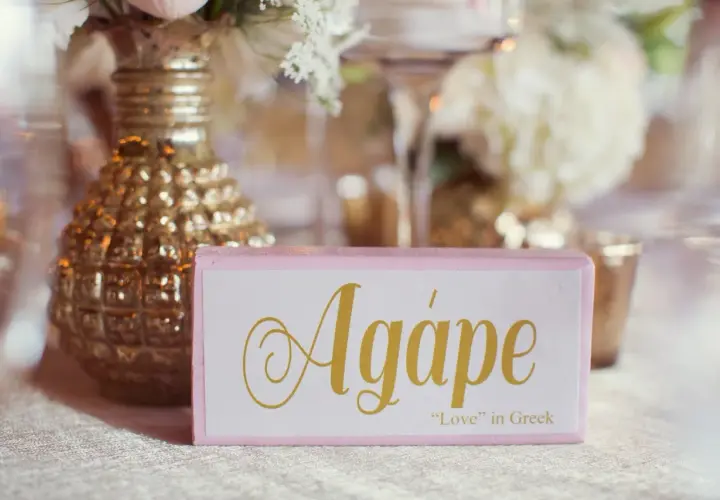 Cartel rosa con la palabra "Agápe" en dorado, junto a un jarrón dorado y flores.