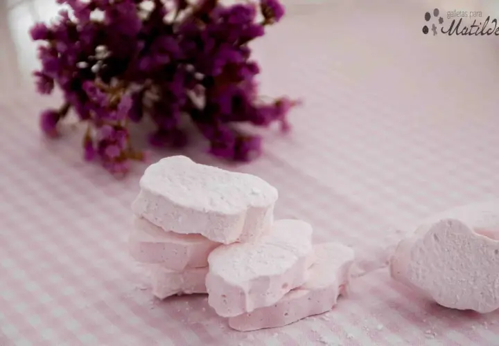 Delicados nubes dulces rosados, espolvoreados con azúcar glas, junto a un ramo de flores moradas sobre un mantel de cuadros.