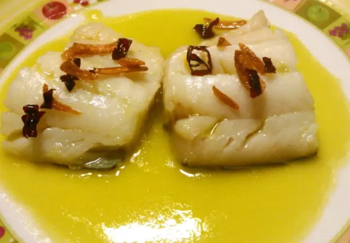 Dos trozos de bacalao al pil pil, cubiertos con aceite de oliva y adornados con ajo frito y guindilla.