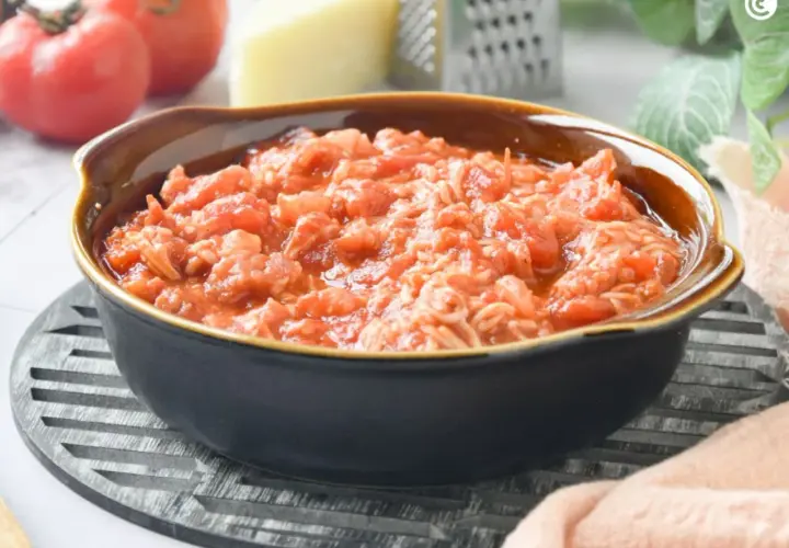 Un tazón de pasta amatriciana, con tomates frescos, queso y rallador al fondo.