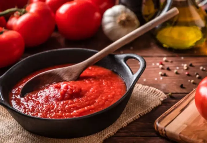 Un tazón de tomate frito casero con una cuchara de madera, rodeado de tomates frescos, ajo y aceite de oliva.