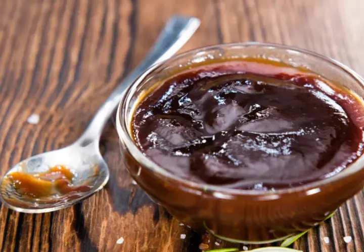 Un cuenco de cristal lleno de deliciosa salsa barbacoa casera, con una cuchara al lado.