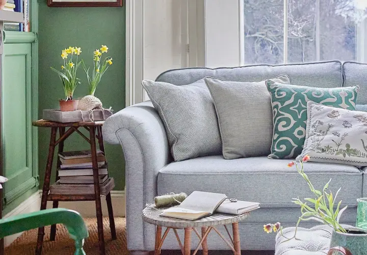 Sofá azul claro con cojines decorativos. Una mesa auxiliar con narcisos y libros complementa la decoración color pastel.