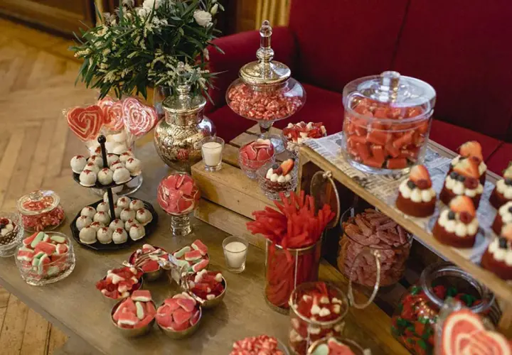Ideas para como hacer un candy bar para boda: mesa llena de dulces rojos y blancos, pasteles, caramelos y piruletas en forma de corazón.