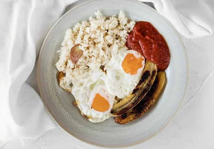 Un plato de arroz a la cubana con dos huevos fritos, plátano maduro frito y salsa de tomate.