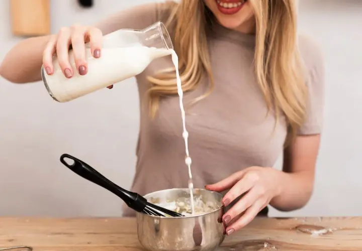 Mujer rubia sonríe mientras vierte leche de una botella a un bol con batidor, preparando algo delicioso.