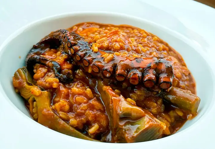 Un tentáculo de pulpo asado reposa sobre un arroz meloso y sabroso, con trozos de alcachofa. Un delicioso arroz con pulpo.