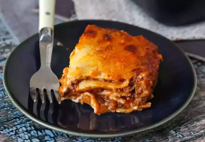 Un trozo de lasaña boloñesa, con capas de pasta, carne y salsa, servido en un plato azul con un tenedor.