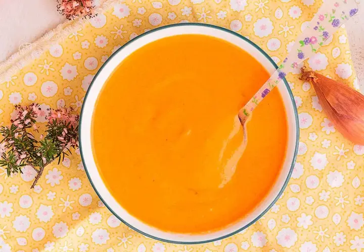 Un tazón de cremosa salsa americana, lista para disfrutar. Una cuchara con flores y una chalota completan la escena.