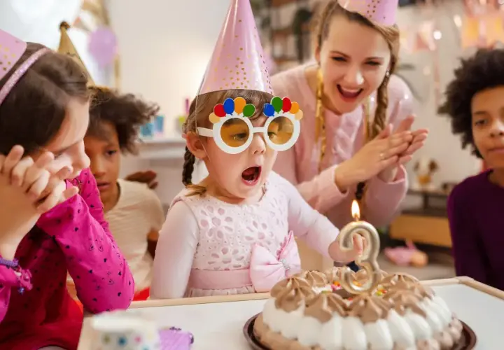 Niños celebran un cumpleaños con pastel y velas. La cumpleañera, con gafas divertidas, sopla la vela. Una temática de cumpleaños para niños llena de alegría.